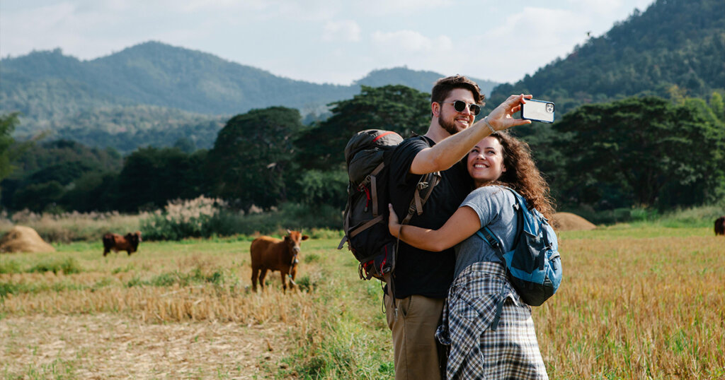 Happy couple of travelers hugging and taking selfie in field against hills