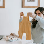 Young woman folding baby clothes sitting near table at home