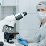 Woman in Protective Clothing Sitting Behind a Microscope