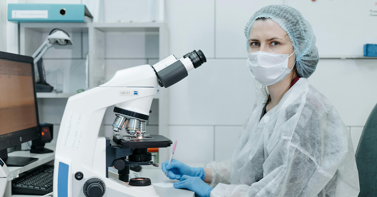 Woman in Protective Clothing Sitting Behind a Microscope